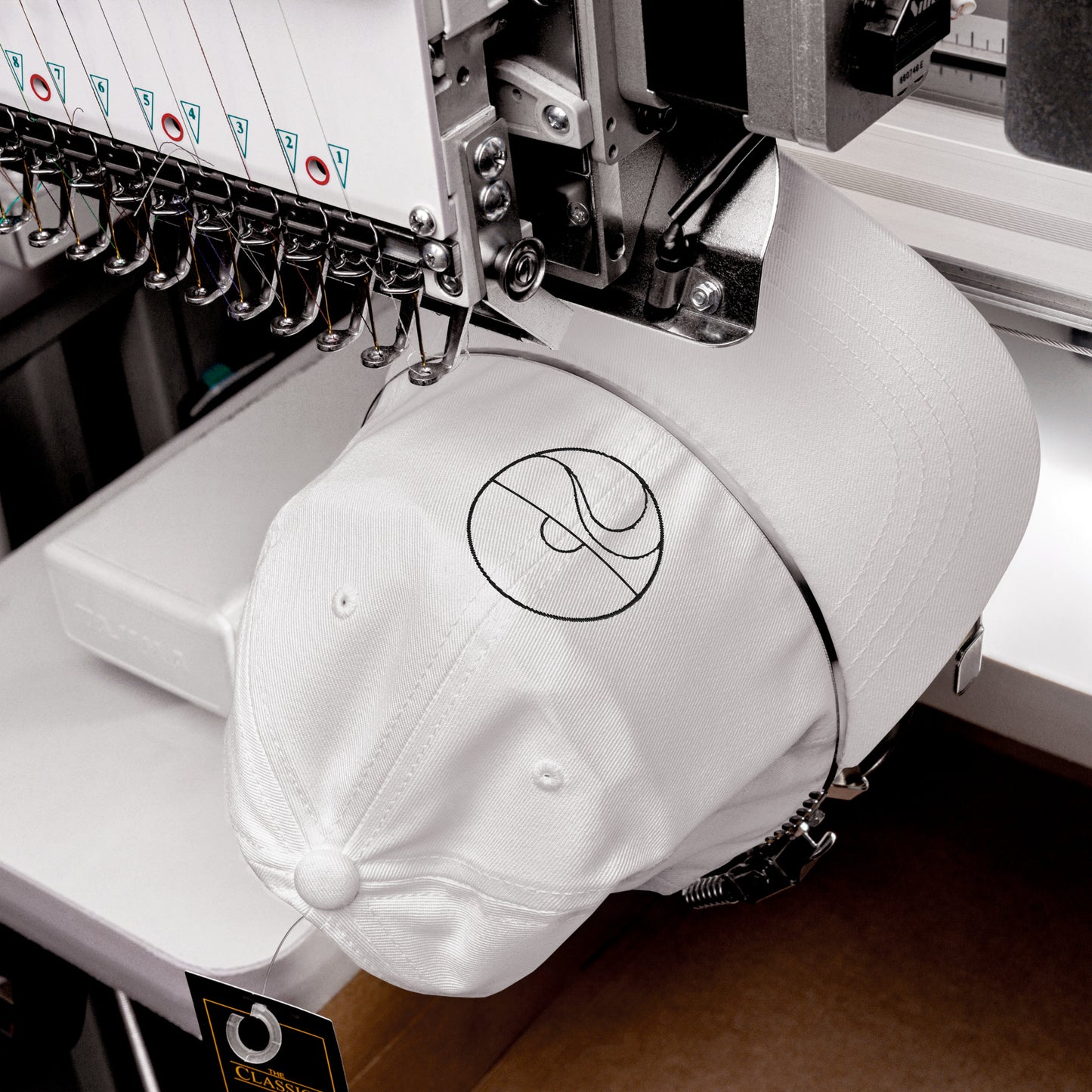 Primer plano de una máquina industrial bordando con hilo negro el diseño de sol y mar sobre una gorra blanca.