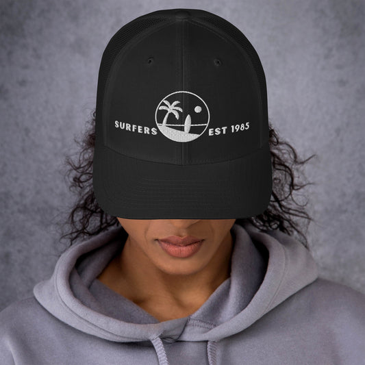 Mujer vistiendo gorra trucker negra estructurada con gráfico de surf de estilo vintage.