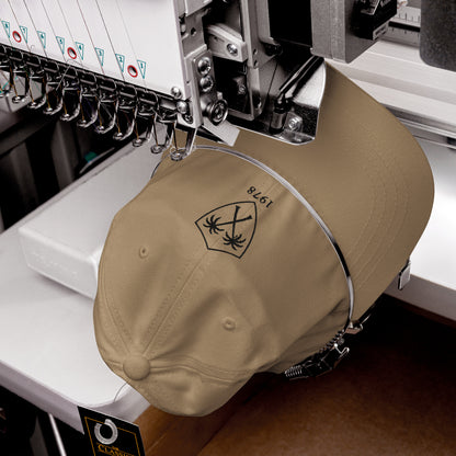 Primer plano de maquinaria industrial realizando el bordado de alta precisión del escudo sobre una gorra caqui.