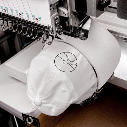 Primer plano de una máquina industrial bordando con hilo negro el diseño de sol y mar sobre una gorra blanca.