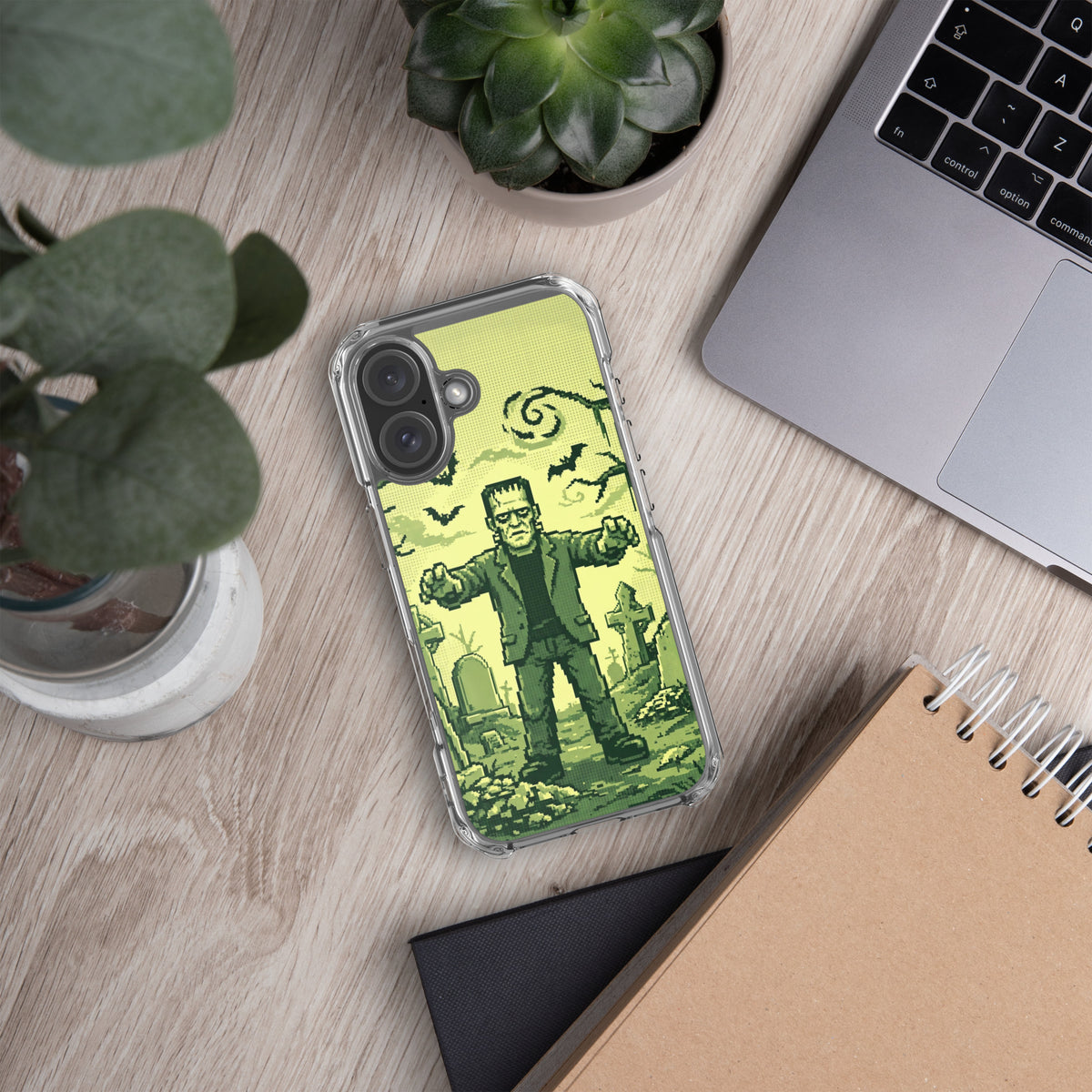 Funda transparente para iPhone® con diseño de Frankenstein en tonos verdes sobre un escritorio junto a plantas y un portátil.