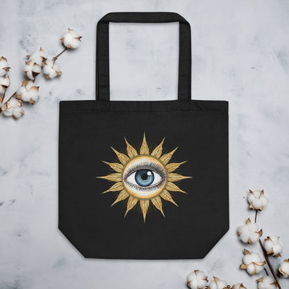 Bolsa de tela negra con diseño de sol místico apoyada sobre una superficie plana junto a elementos decorativos botánicos.