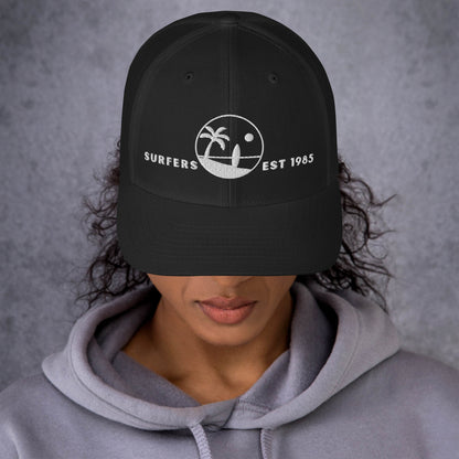 Mujer vistiendo gorra trucker negra estructurada con gráfico de surf de estilo vintage.