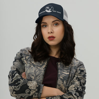 Mujer vistiendo gorra trucker azul marino y blanca con ilustración clásica de palmera y tabla de surf.